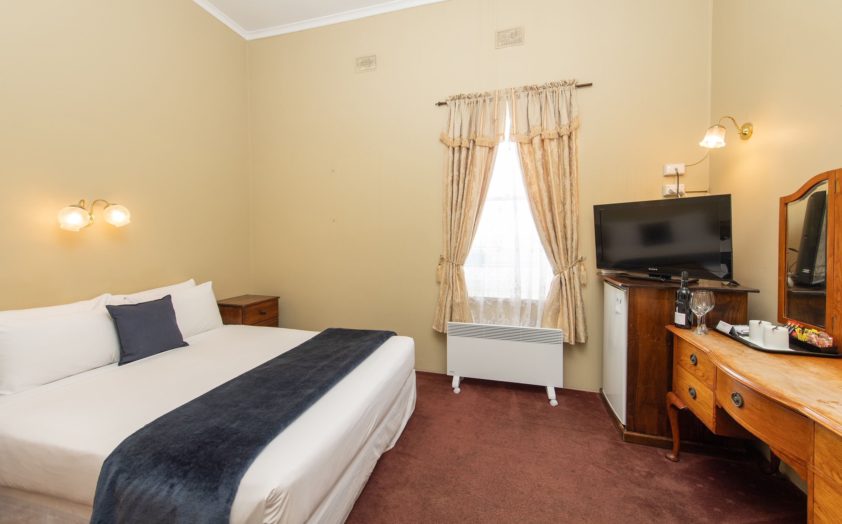 A hotel room with a bed, a desk with a television, a dresser, and a window with curtains.