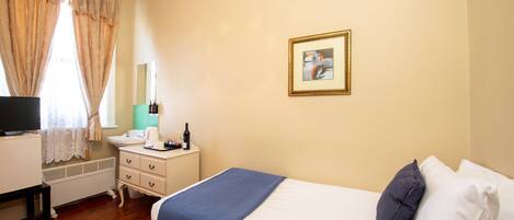 A hotel room with a bed, a bedside table with a lamp, a small desk with a computer, a window with curtains, and a framed picture on the wall.