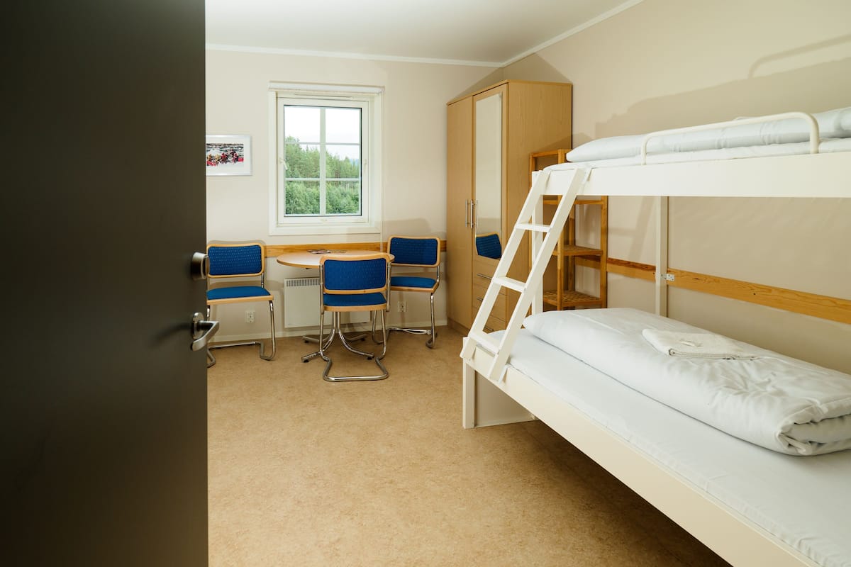 A bunk bed room with a desk and chairs, a window, and a wardrobe.