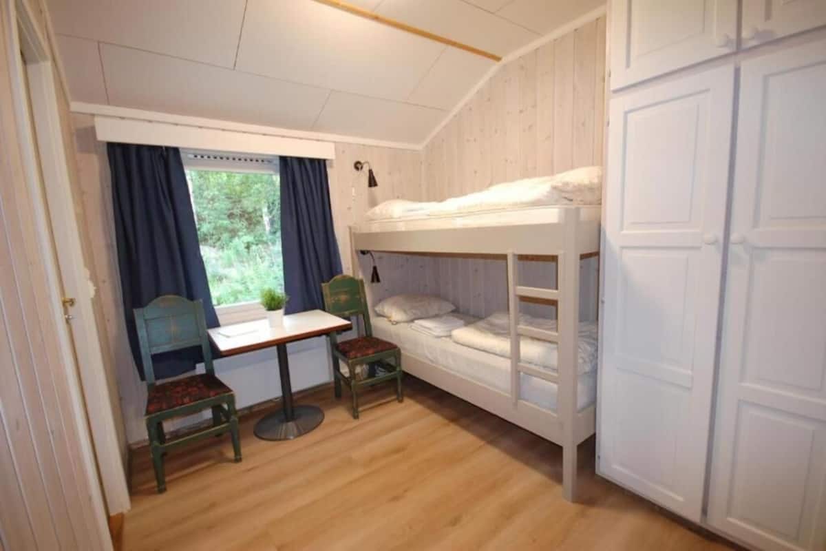 A small, cozy room with a bunk bed, a desk, two chairs, and a window with curtains.