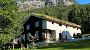A two-story log cabin with a green roof, surrounded by a grassy area and a mountain backdrop.