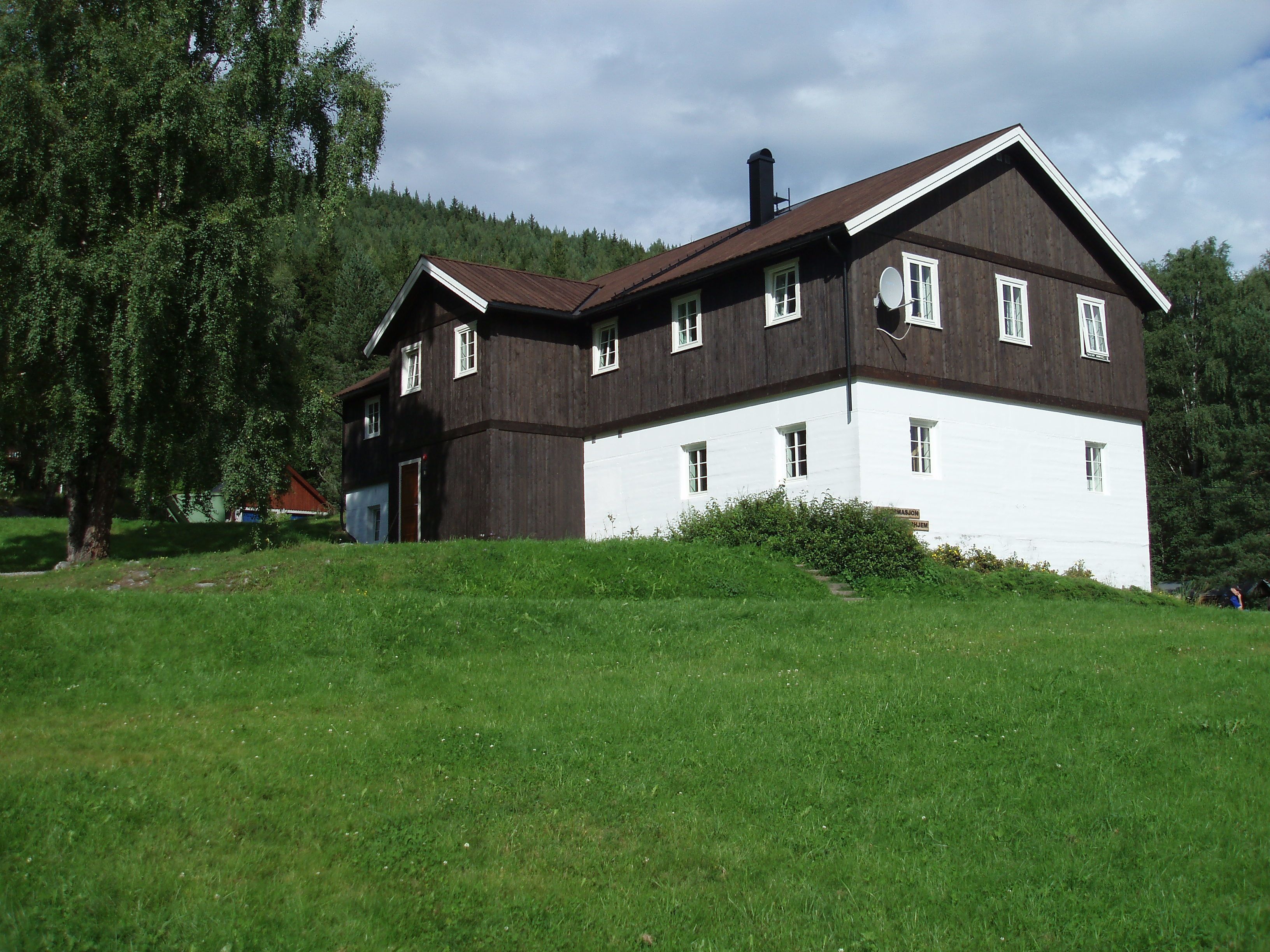A two-story wooden house with a white lower section and a brown upper section, surrounded by greenery and trees.