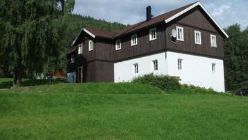 A two-story wooden house with a white lower section and a brown upper section, surrounded by greenery and trees.