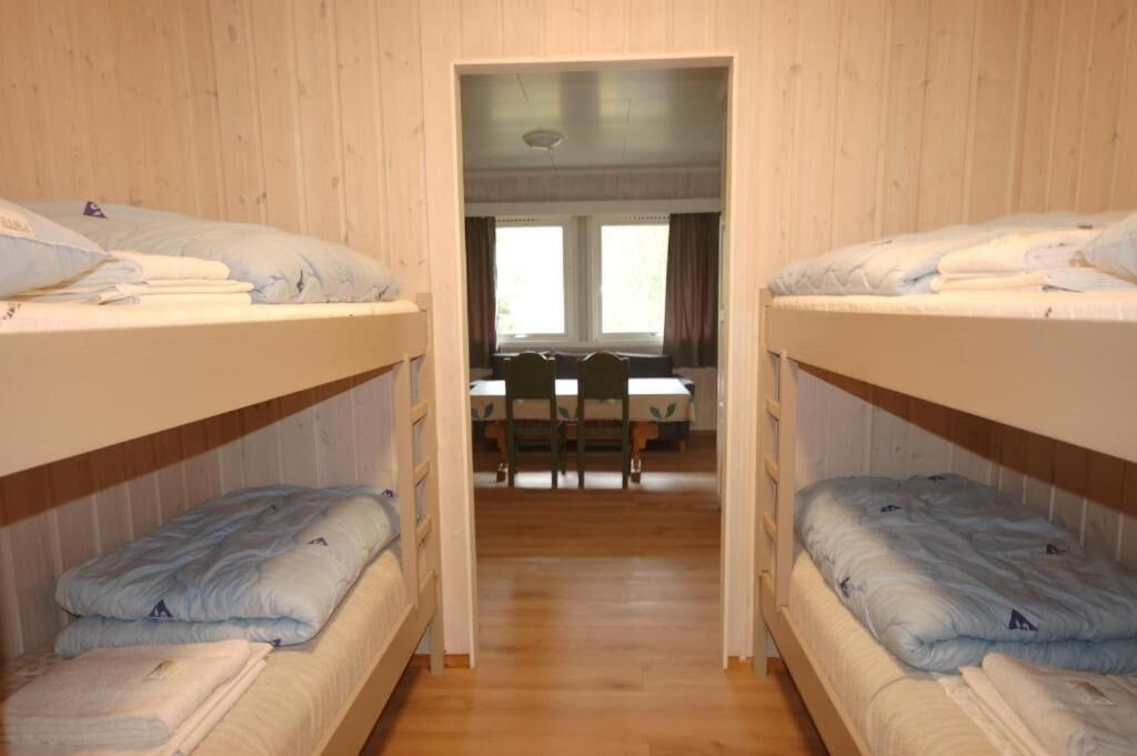 Quarto com beliches, piso de madeira, mesa com cadeiras e janela com cortinas.