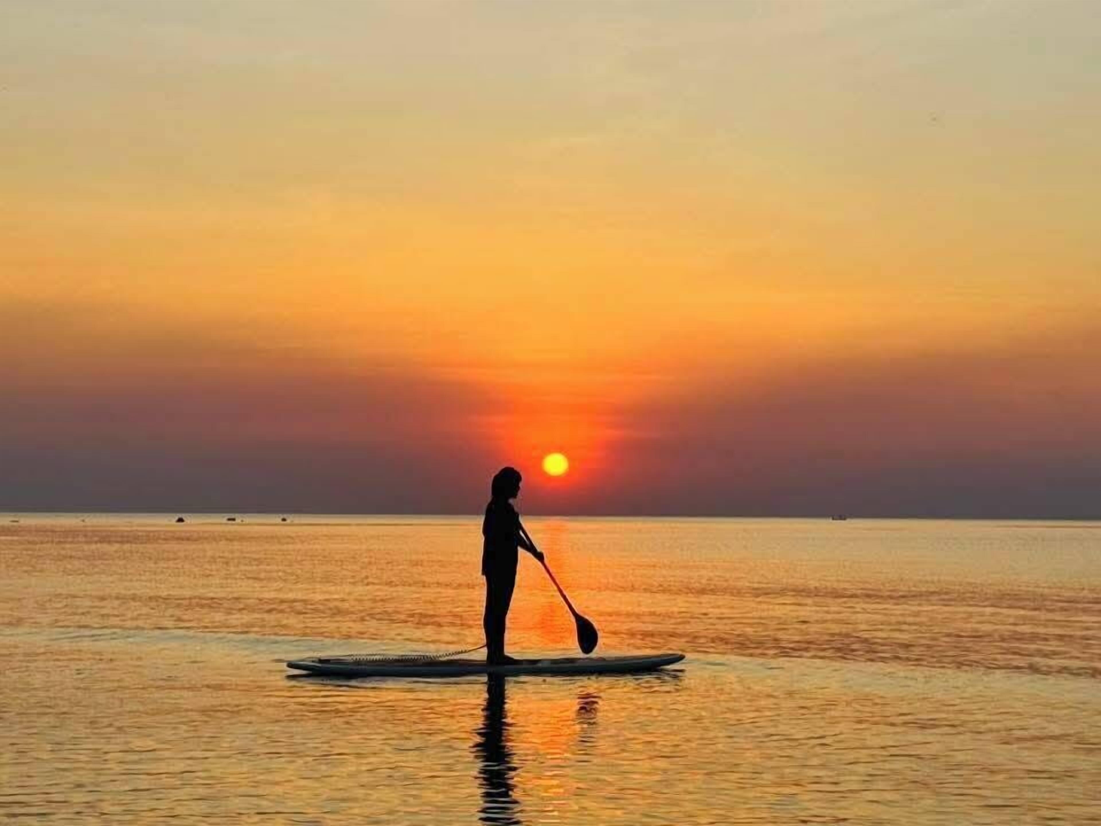 Une personne pratiquant le paddle au coucher du soleil, avec le soleil visible en arrière-plan.