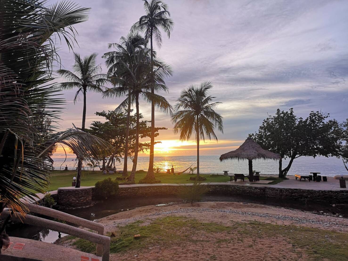 Vista de la playa con palmeras, una estructura de techo de paja y un atardecer sobre el océano.