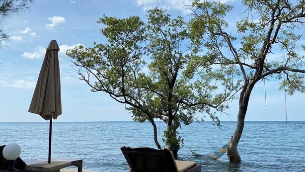 Une terrasse en bord de mer avec une table en béton, un fauteuil de jardin en bois et un parasol de plage.