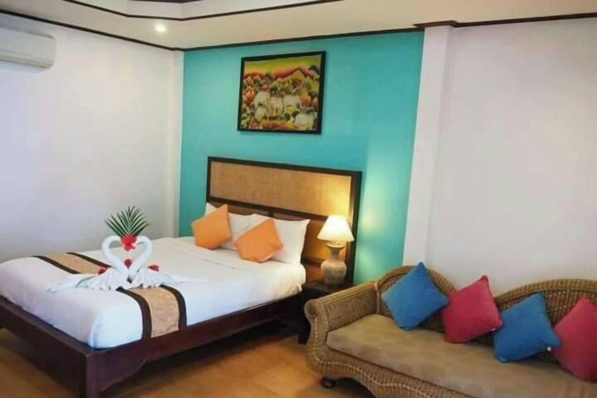 A hotel room with a bed, a nightstand with a lamp, a sofa with cushions, and a painting on the wall.
