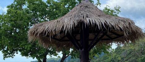 A beachside lounge with a thatched umbrella, wooden lounge chairs, and a view of the sea and greenery.