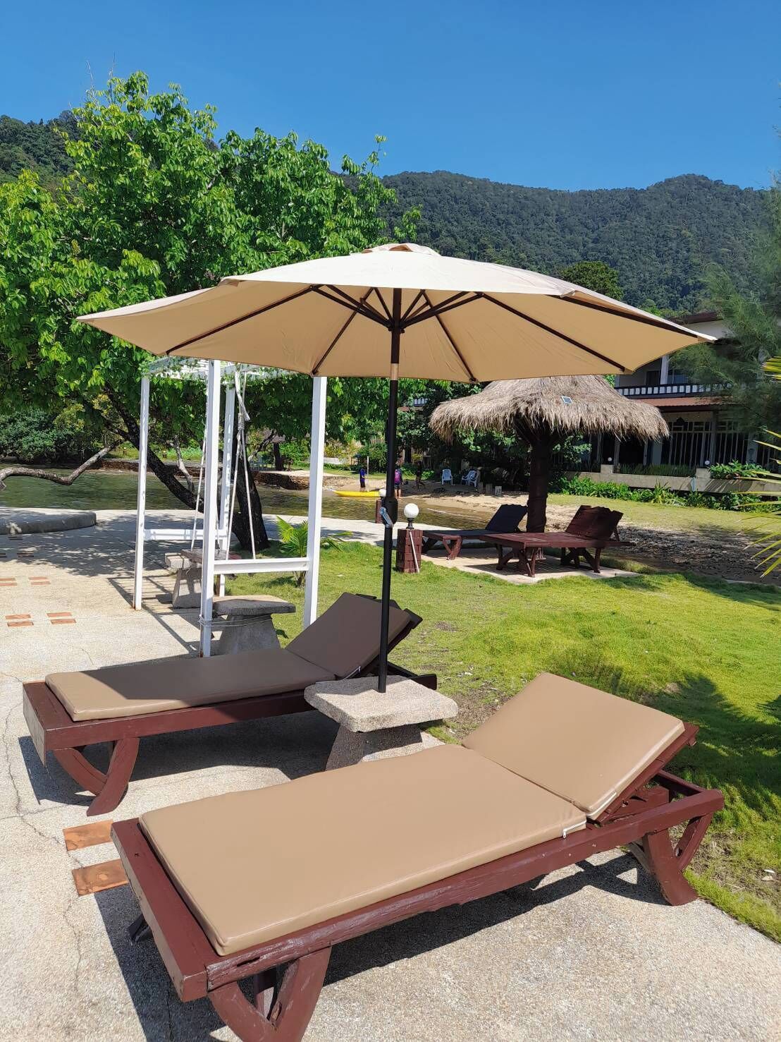 A poolside area with lounge chairs and a large umbrella, surrounded by greenery and a mountain in the background.