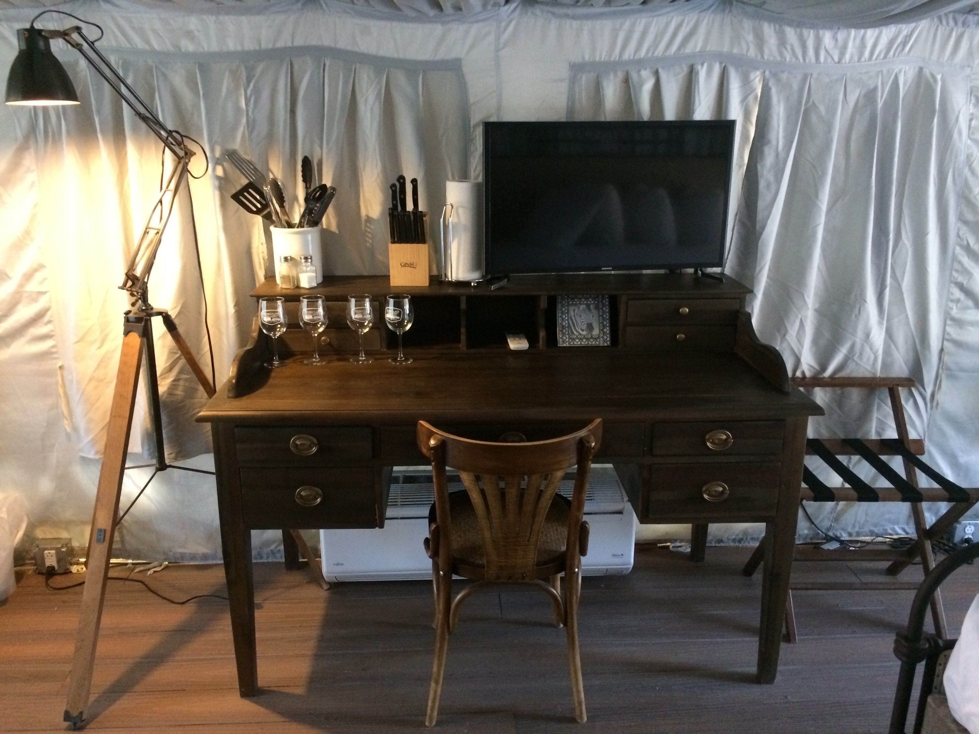 A wooden desk with a television, a chair, and a lamp. There are wine glasses and a knife block on the desk.