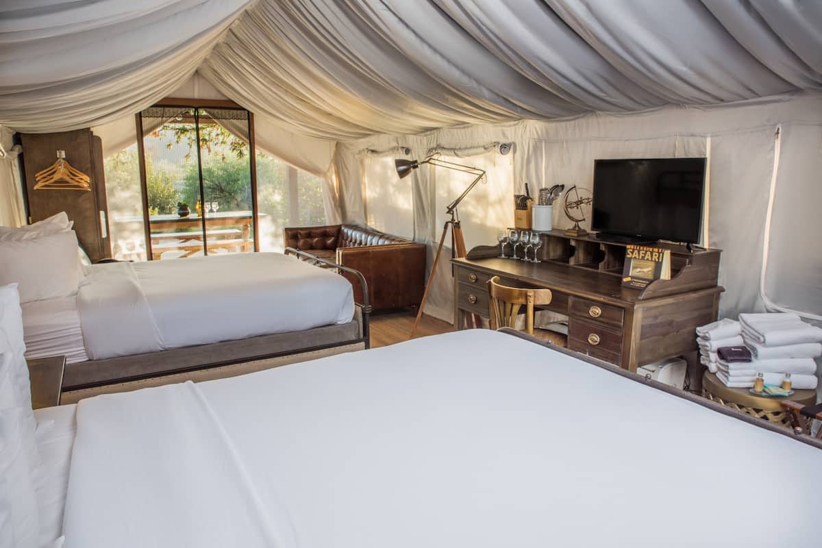 A tented room with two beds, a wooden dresser, a television, and a window with a view of greenery.