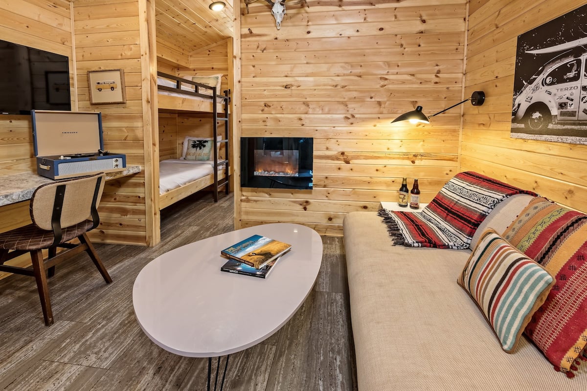 A cozy cabin with a bunk bed, a fireplace, a sofa with patterned cushions, and a small table with books.