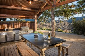 An outdoor kitchen with a grill, a table with wine glasses and a bottle, and a view of the surrounding landscape.