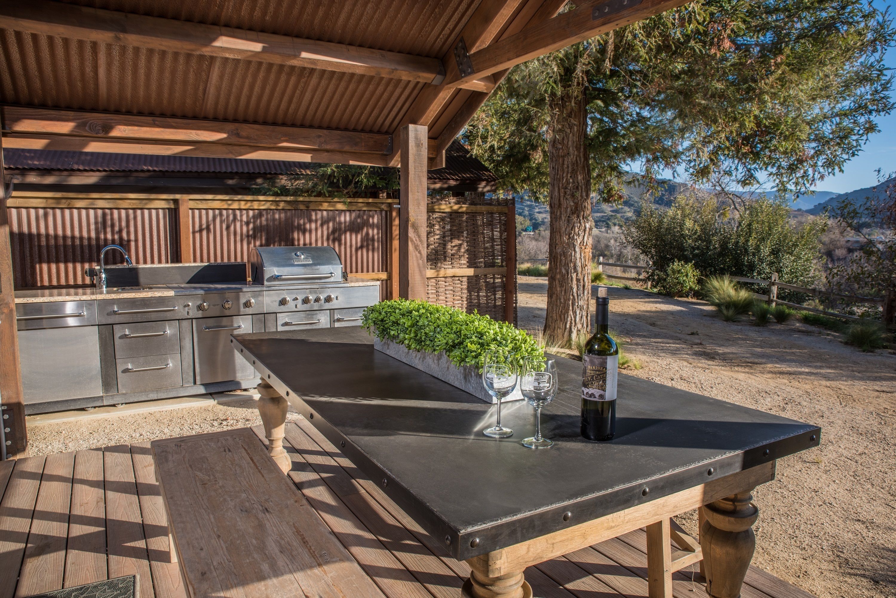 Eine Außenküche mit Grill, Spüle und Edelstahlgeräten, ein Holztisch mit Flasche und Gläsern sowie ein Blick auf die umliegende Landschaft.