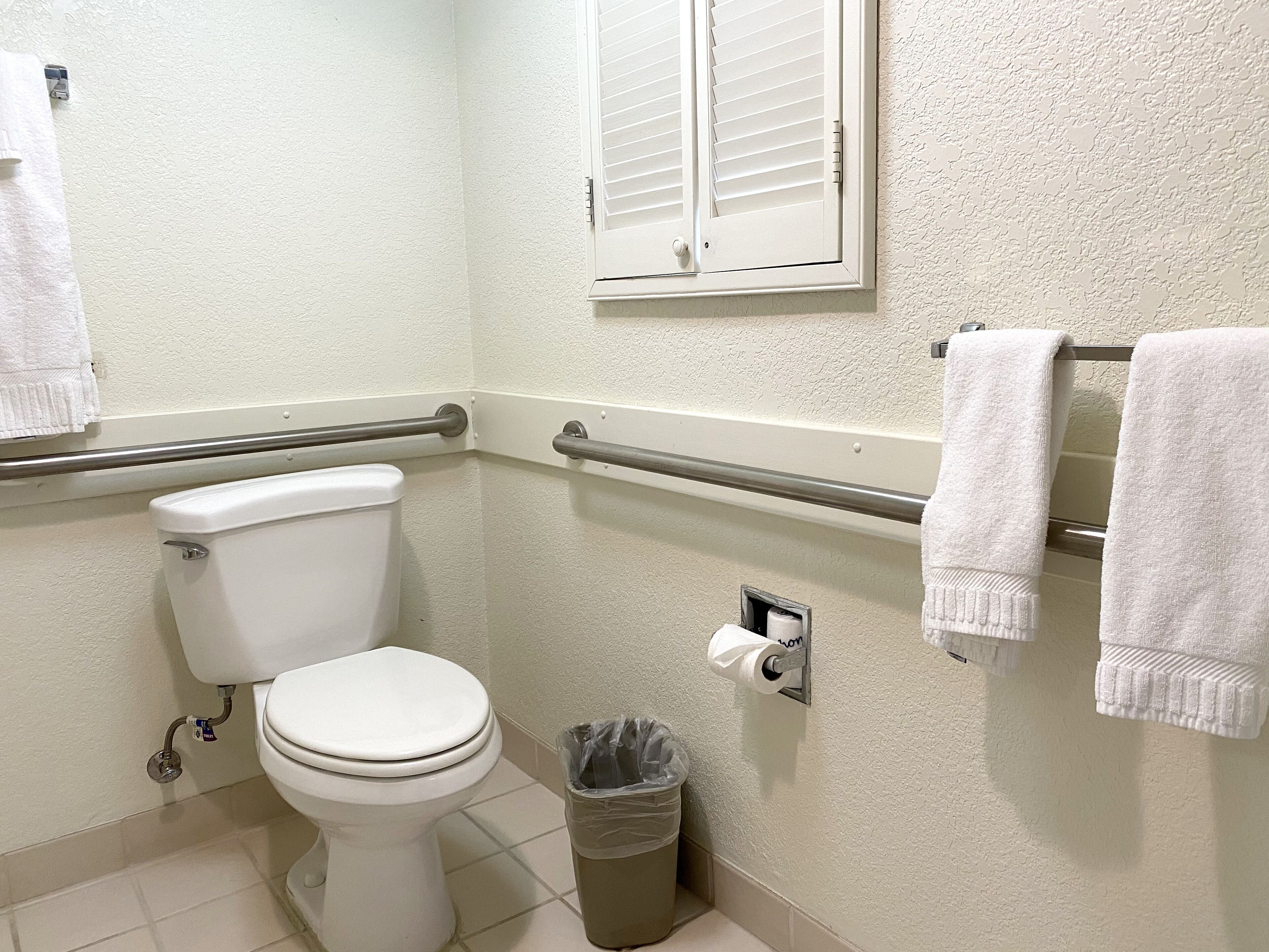 Ein Badezimmer mit Toilette, Haltegriffen, einem Mülleimer, einem Handtuchhalter und einem Fenster mit Jalousien.