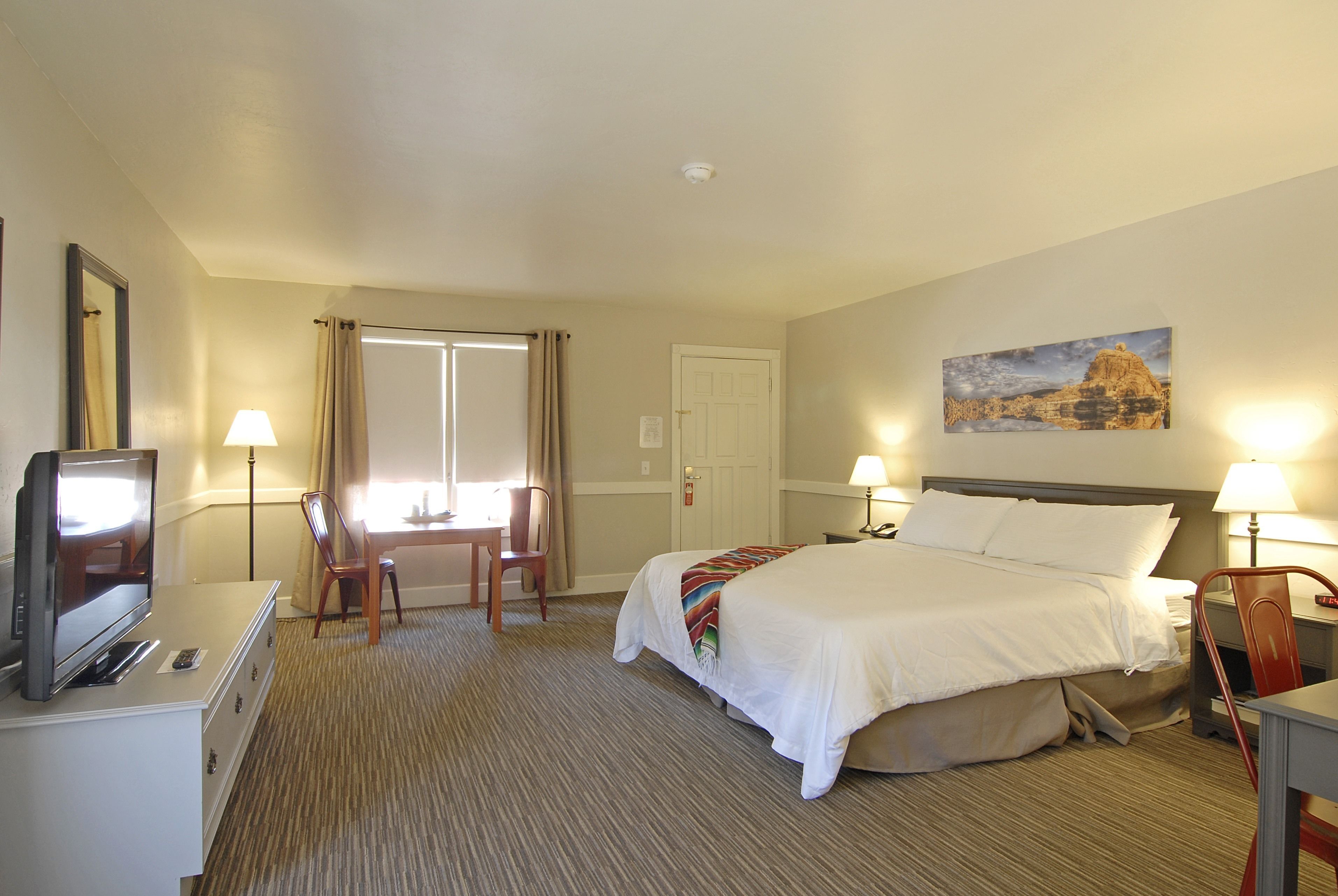 A hotel room with a bed, a desk with a TV, a chair, a lamp, and a painting on the wall.