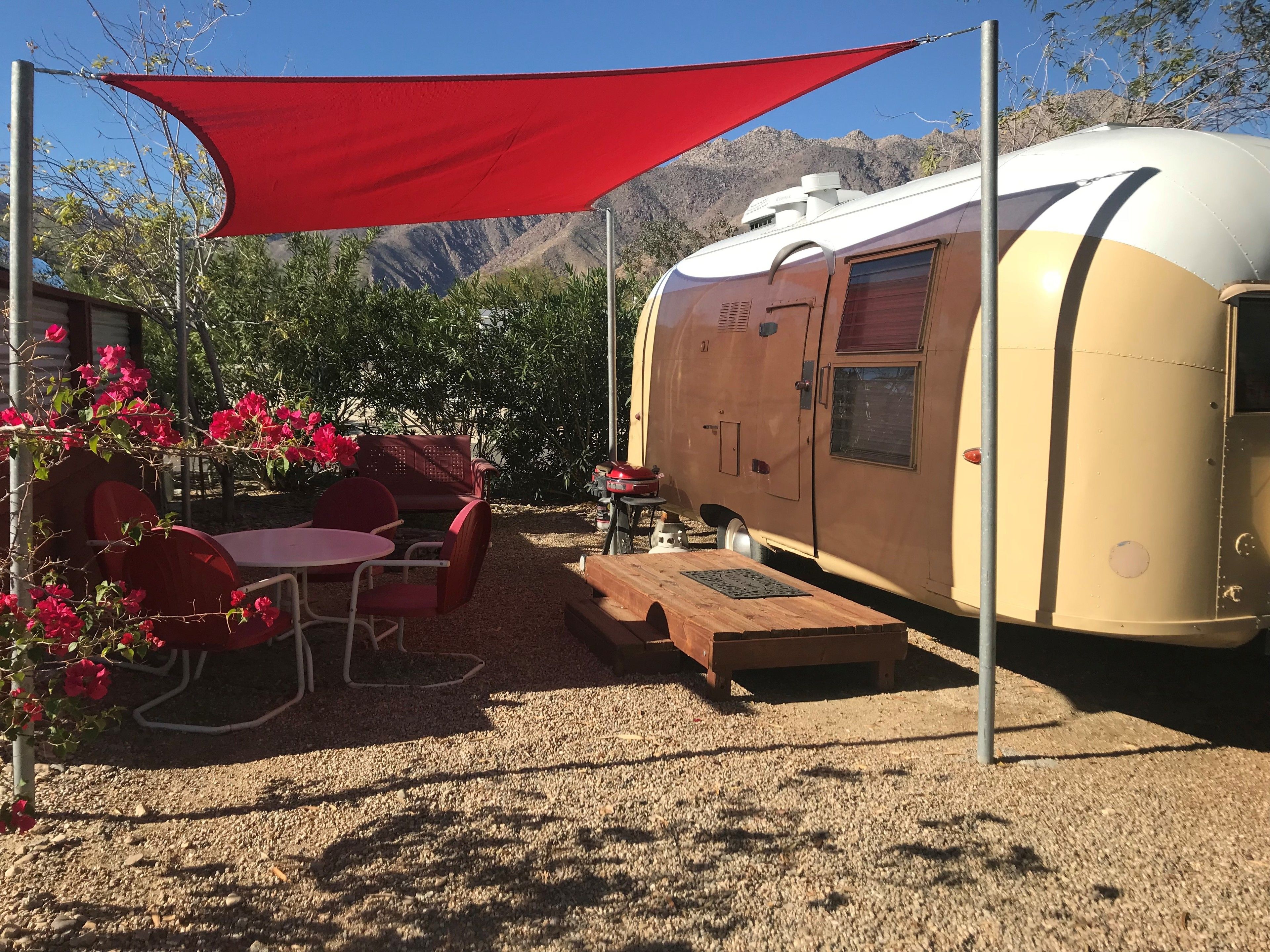 Una caravana con zona de comedor, que incluye una mesa y sillas, instalada en un camping con una vela de protección roja sobre ella.