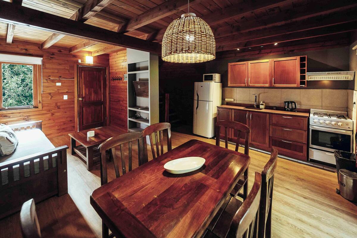 Ein rustikaler Holzinnenraum mit Esstisch, Stühlen, einer Pendelleuchte, Holzschränken, einem Kühlschrank und einem Herd.