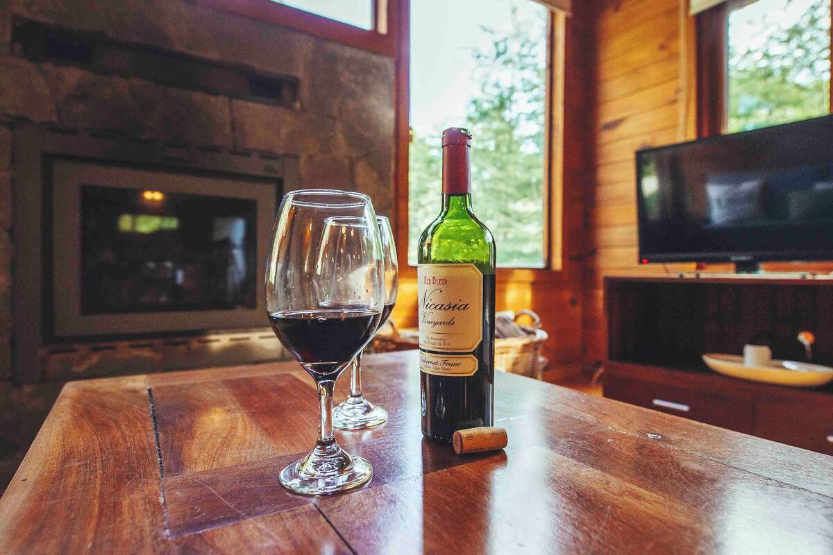 Ein Holztisch mit einer Flasche Rotwein und einem Weinglas, einem Fernseher und einem Kamin in einem rustikalen Ambiente.