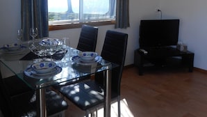 Uma sala de jantar com mesa de vidro, cadeiras e uma televisão sobre um suporte.