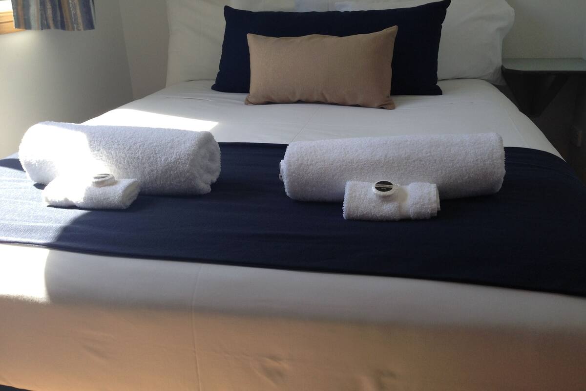 A neatly made bed with white and navy bedding, two rolled towels, and a watch on one of the towels.