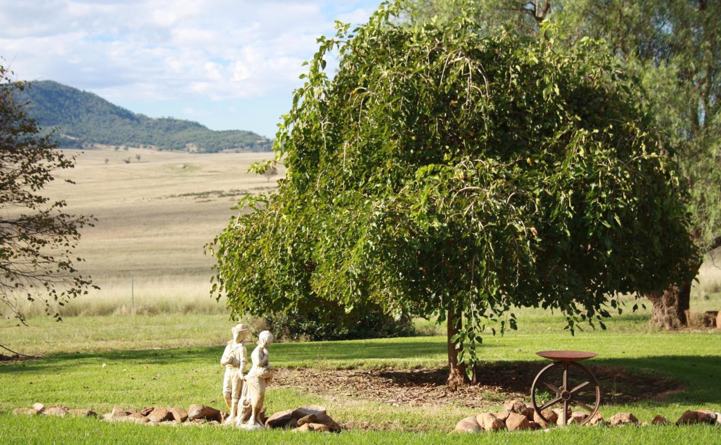 A park with a large tree, a stone statue, and a metal table.