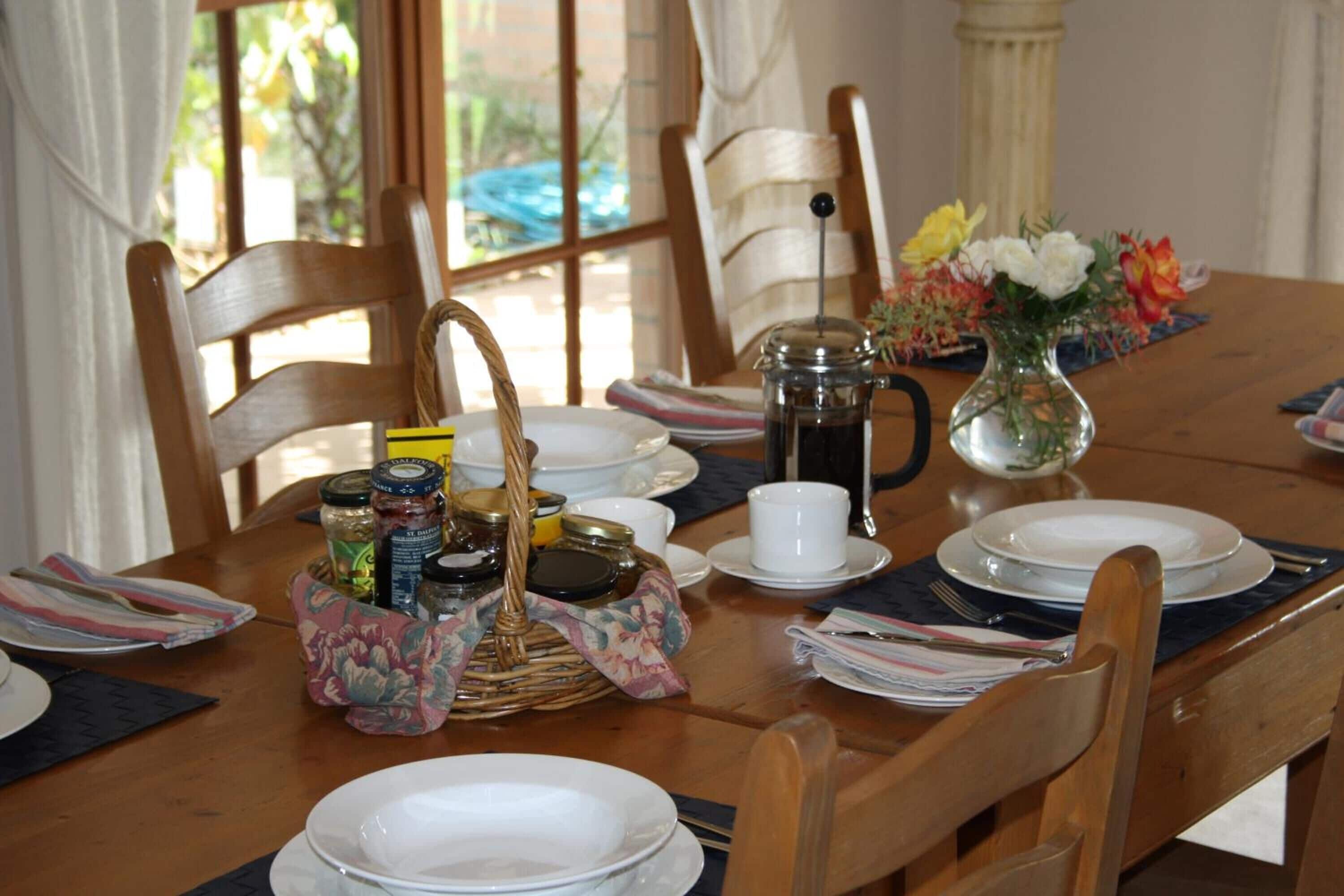A dining table set with plates, a coffee pot, a vase with flowers, and a basket of condiments.