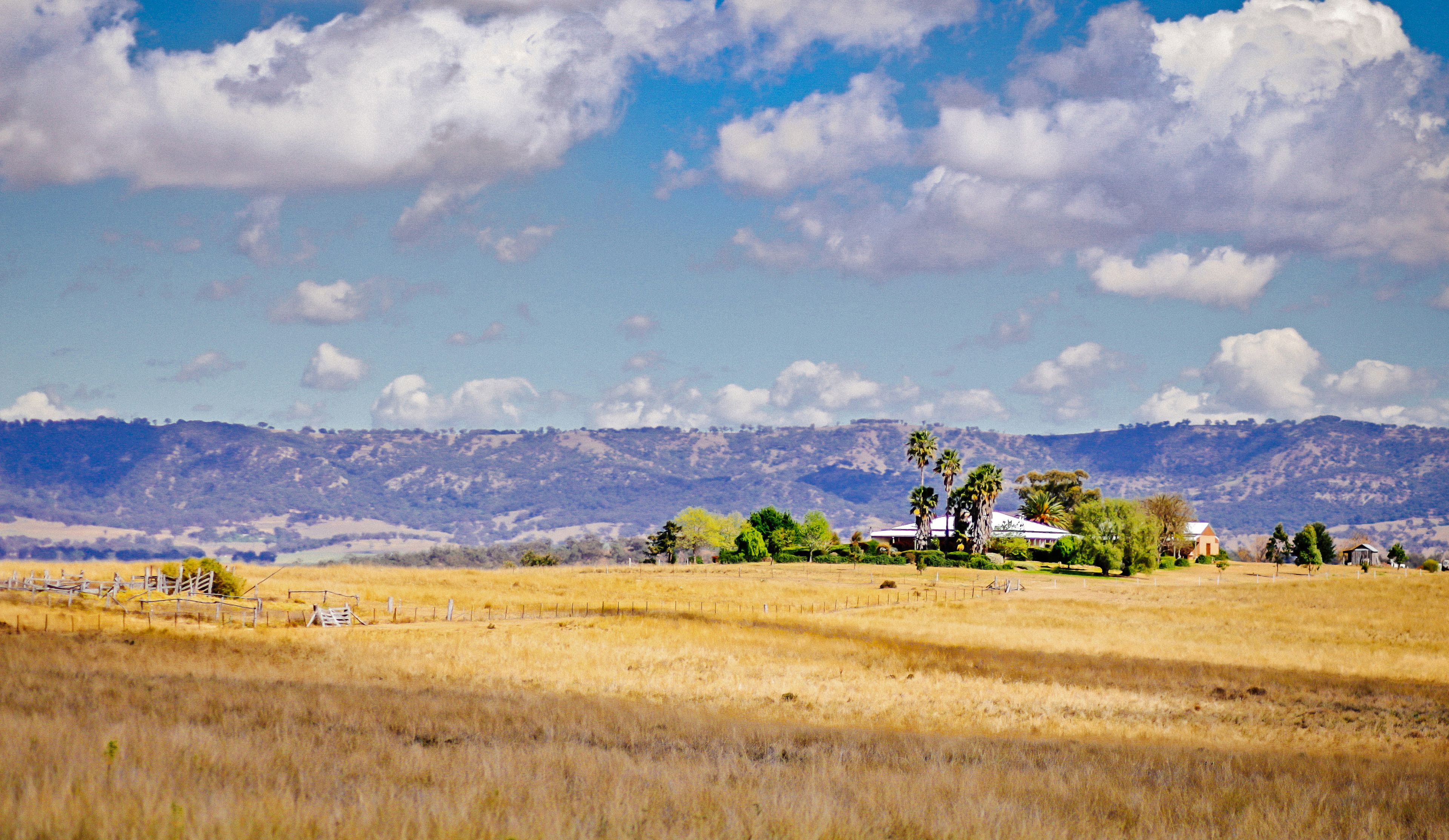 A rural landscape with a house surrounded by fields and mountains in the background.