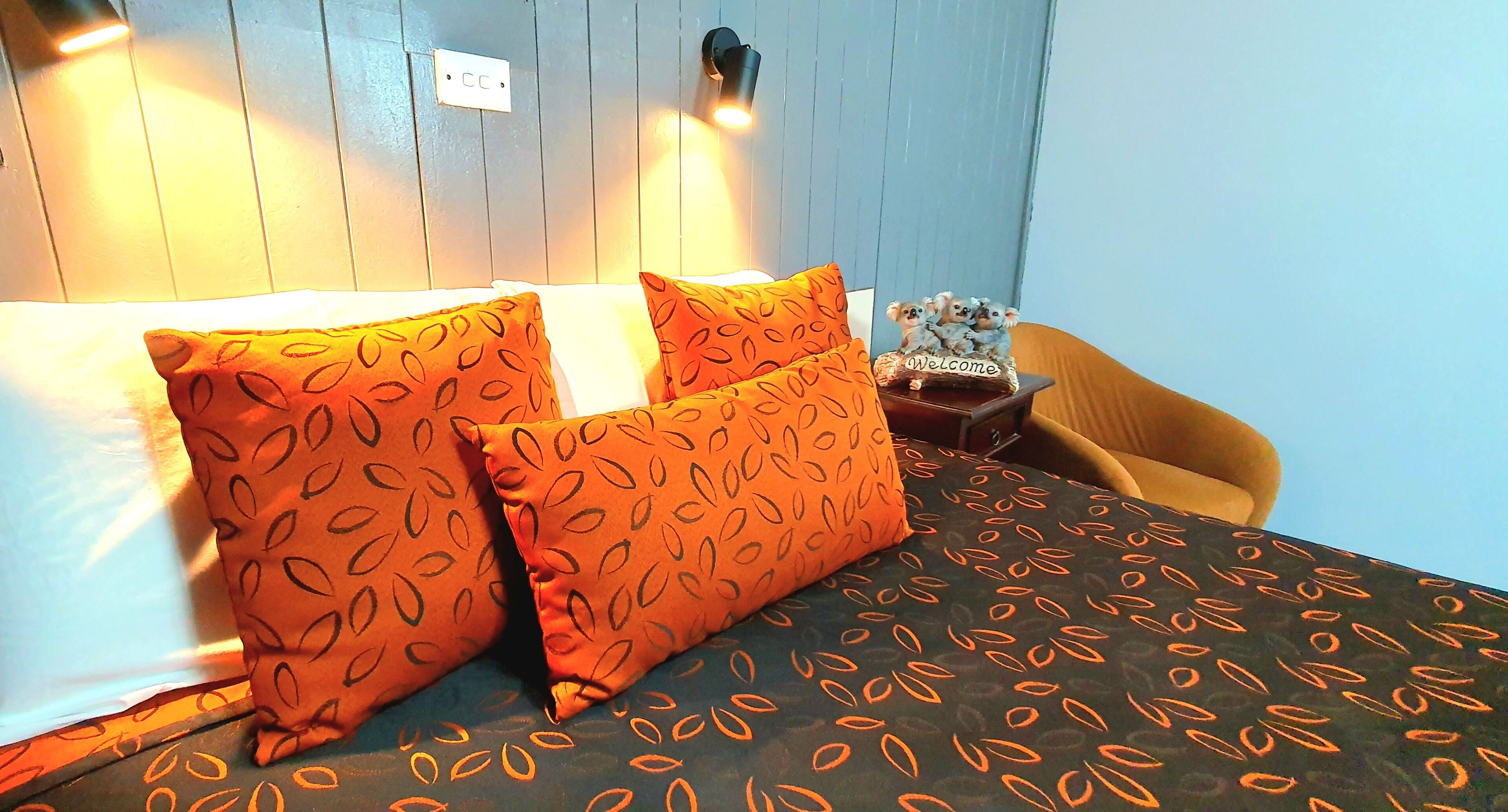 Habitación de hotel con cama, almohadas estampadas color naranja, una mesita con un cartel de 'Welcome' y pared azul.