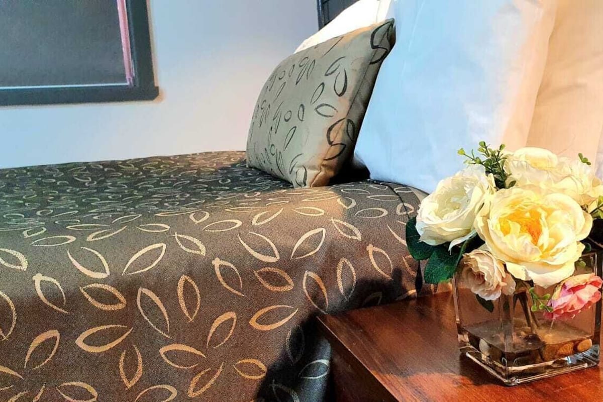 Una cama bien tendida con ropa de cama estampada, una mesita de noche con un jarrón de flores blancas y una lámpara de noche.