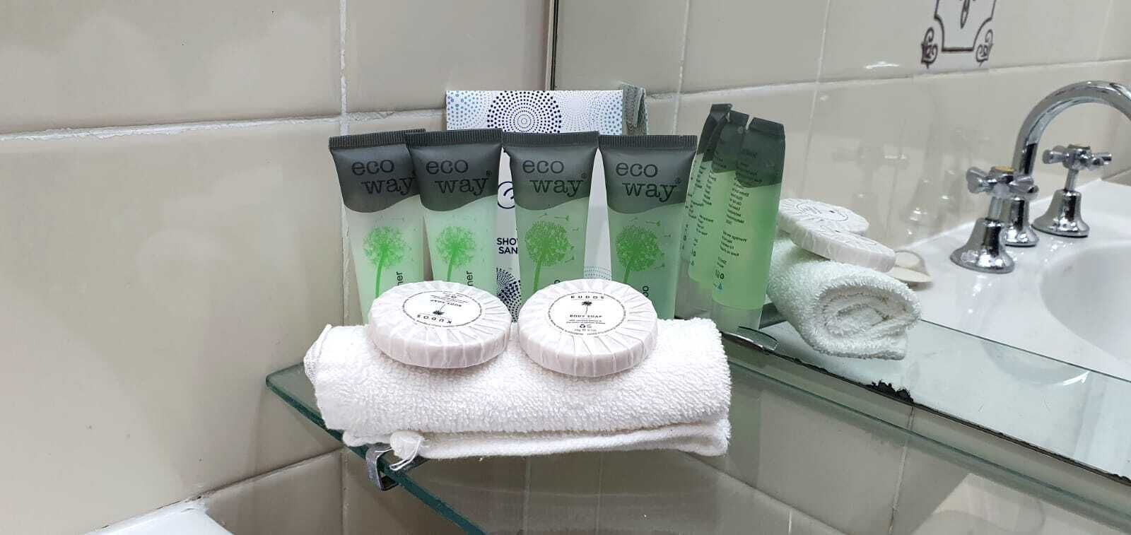 A bathroom with a glass shelf holding a white towel and two white bath mats. Behind the shelf, there are several eco-friendly personal care products.