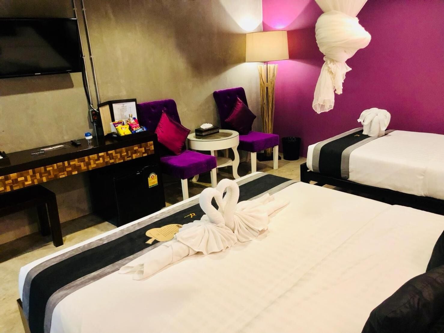 Ein Hotelzimmer mit einem Bett, zwei violetten Ohrensesseln, einem kleinen runden Tisch, einem Schreibtisch mit Computer und Snacks sowie einem Fernseher an der Wand.