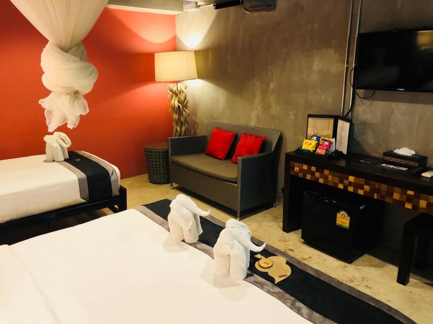 Habitación de hotel con dos camas, un sofá con cojines rojos, un escritorio con televisor y un cesto de basura.