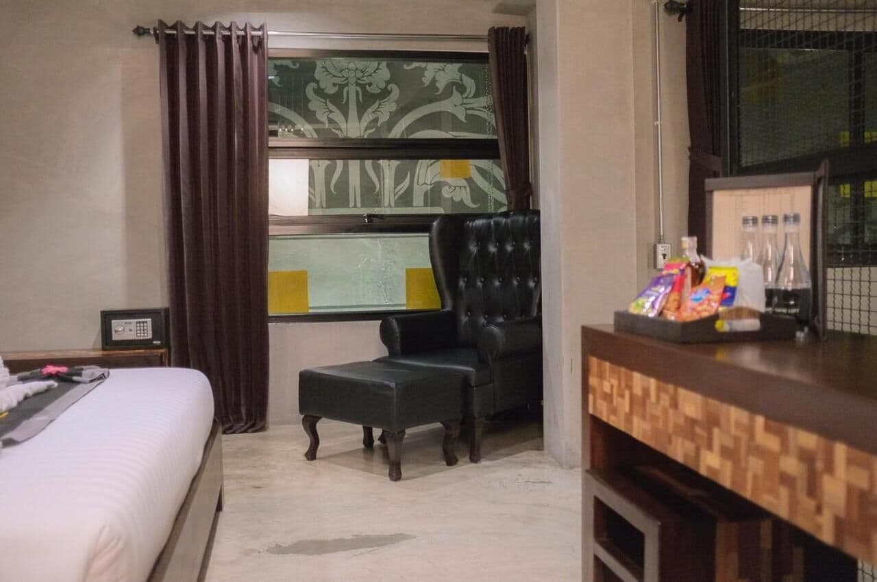 A hotel room with a bed, a black leather armchair, a small wooden table with snacks, a window with curtains, and a wall-mounted phone.