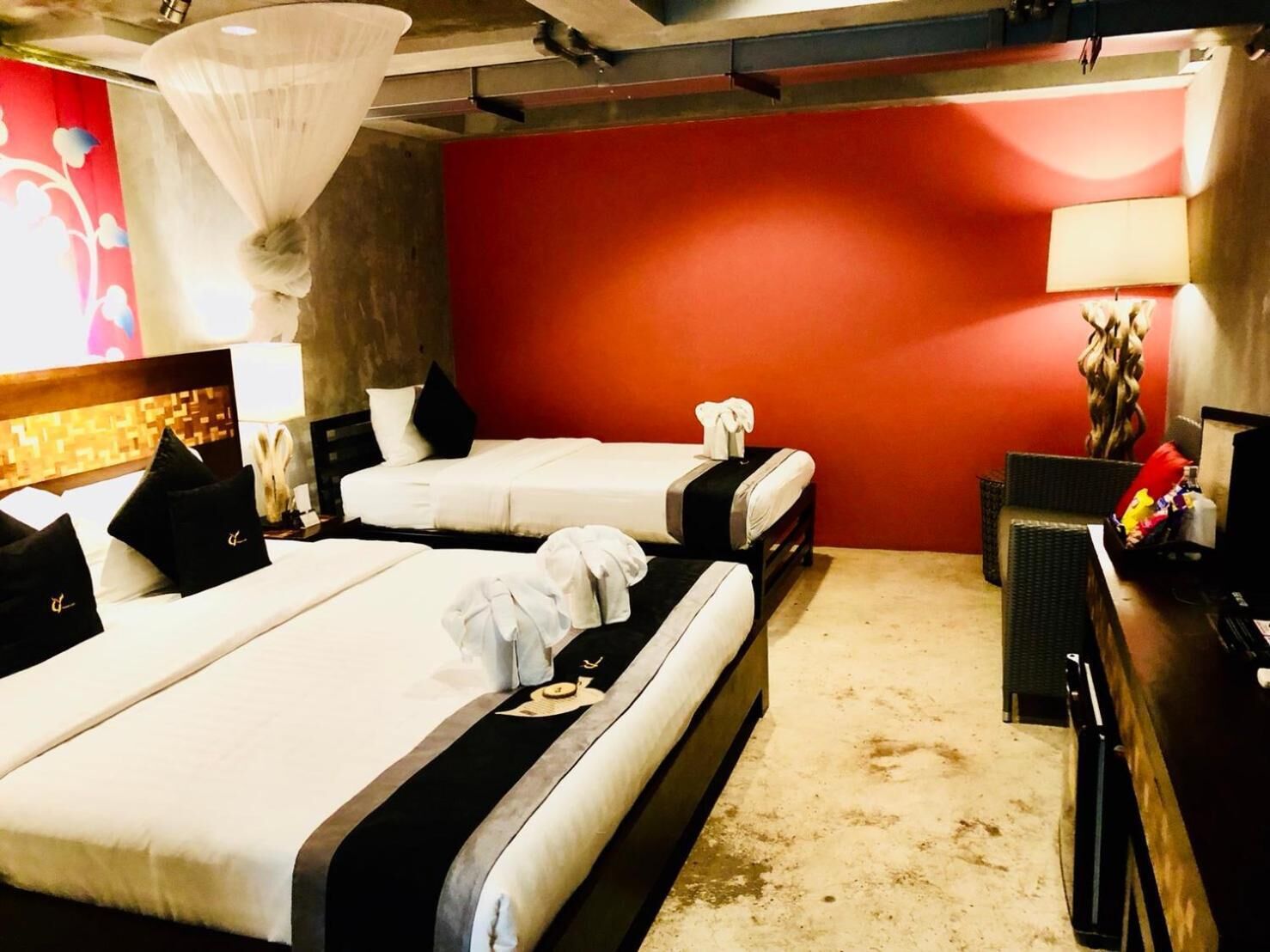 A hotel room with two beds, a red accent wall, a white canopy bed, a desk with a lamp, and a wicker chair.