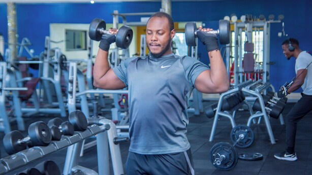 A gym with weightlifting equipment, a person exercising, and another person in the background. - Mika Convention Center Meanwood (Lusaka)