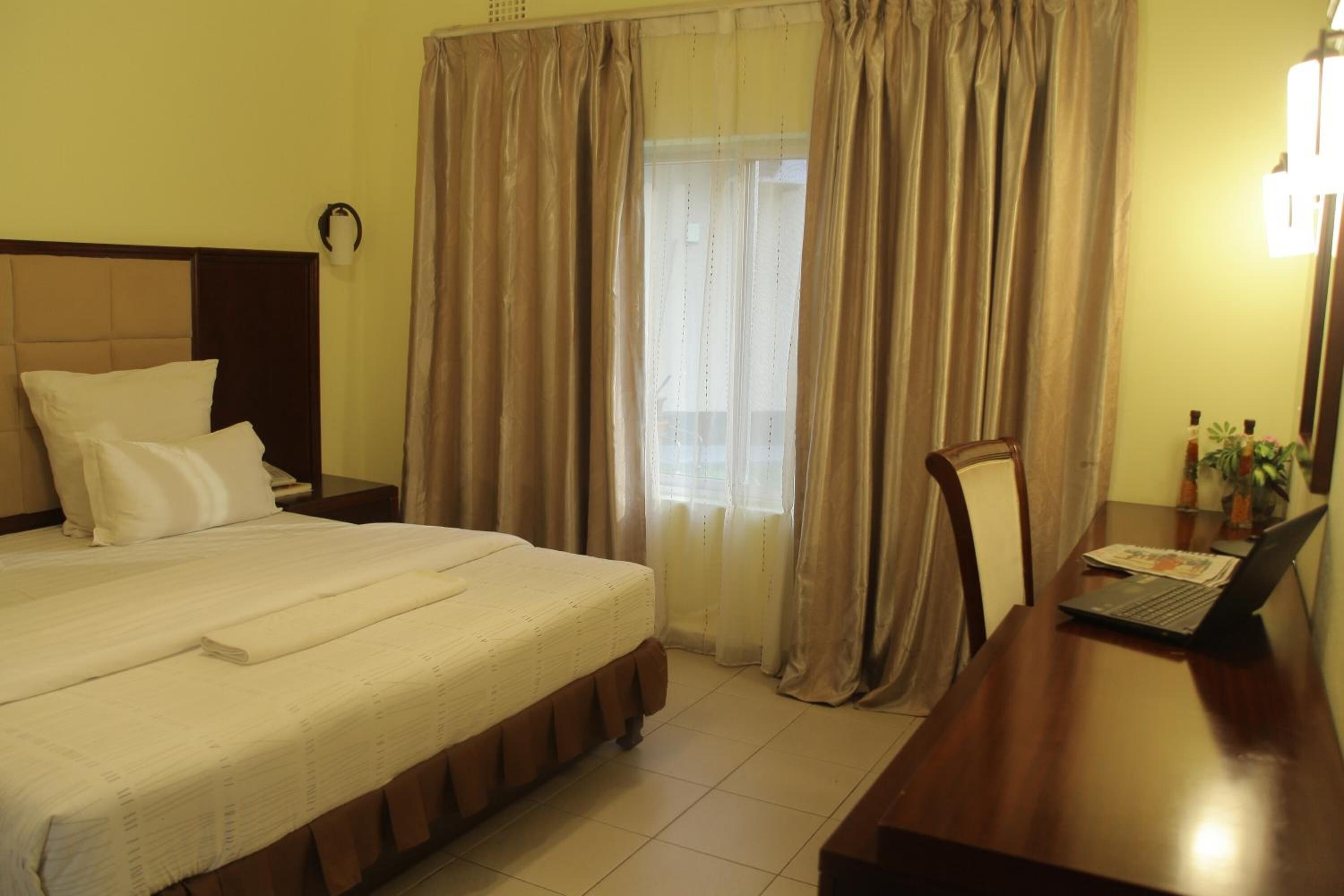 A hotel room with a bed, a desk with a laptop, a chair, a nightstand, and a window with curtains.