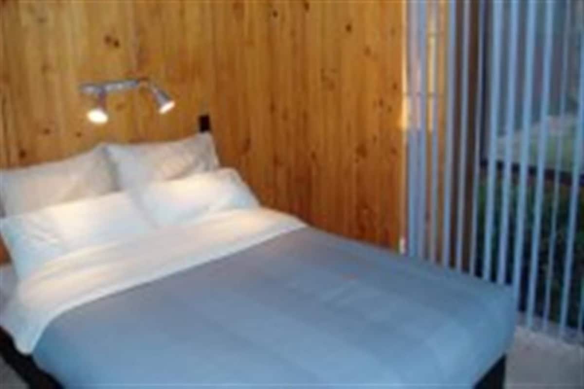 A bedroom with a wooden wall, a bed with white bedding, and a window with vertical blinds.