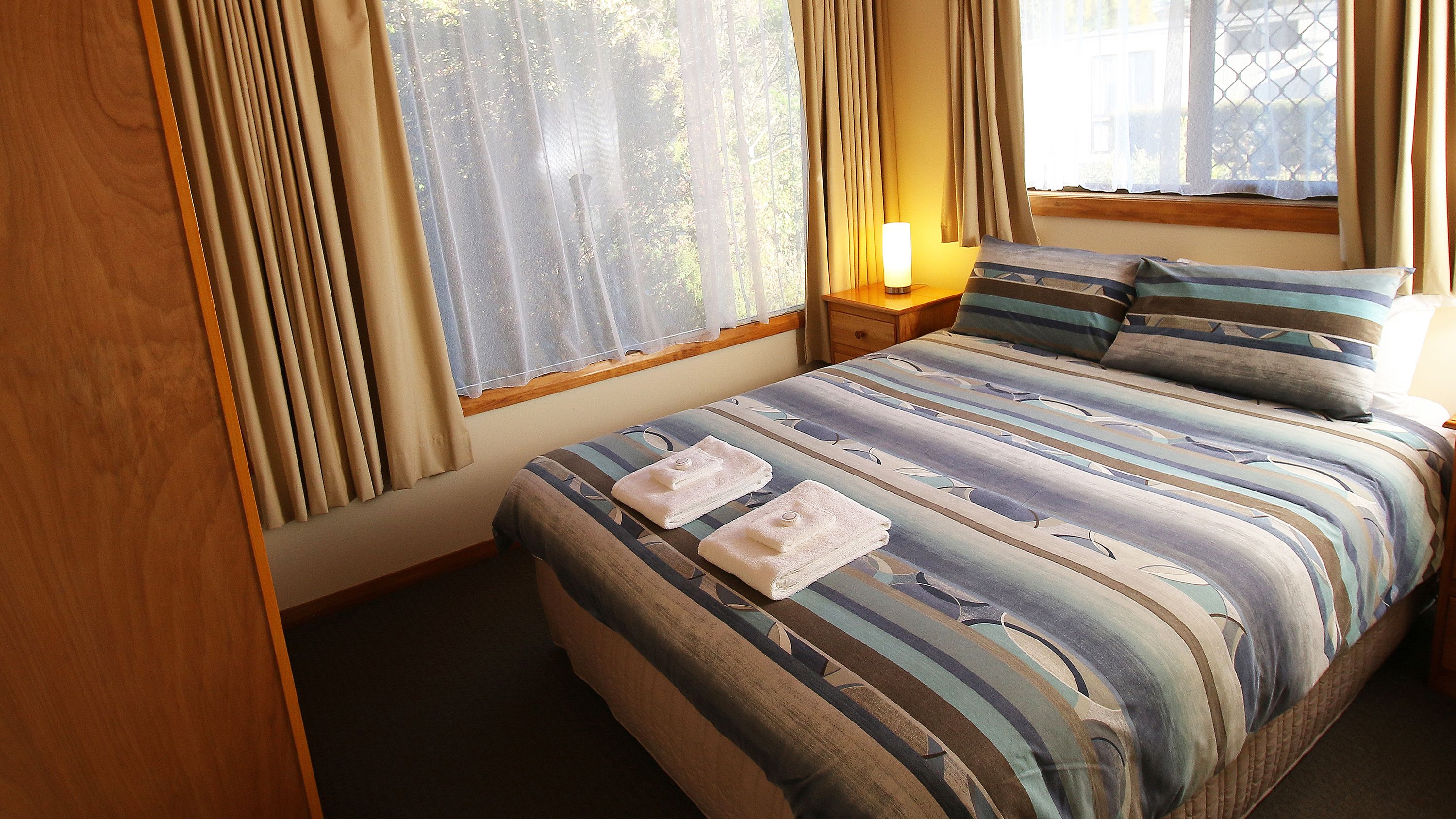 Una cama bien hecha con ropa de cama a rayas y dos toallas, una mesita de noche con lámpara y una ventana con cortinas.
