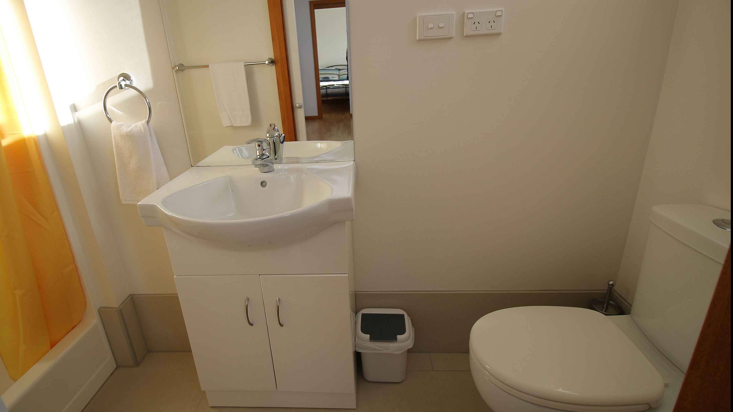 Une petite salle de bain propre, avec un lavabo blanc, un miroir et des toilettes. On peut voir une poubelle et un porte-serviettes.