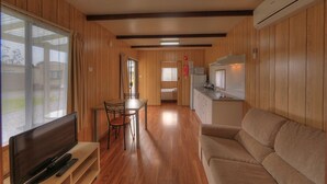 Habitació compacta amb interior de fusta, un sofà, una televisió, una taula de dinar amb cadires, un espai kitchenette i una porta corredissa que dona a zona exterior.