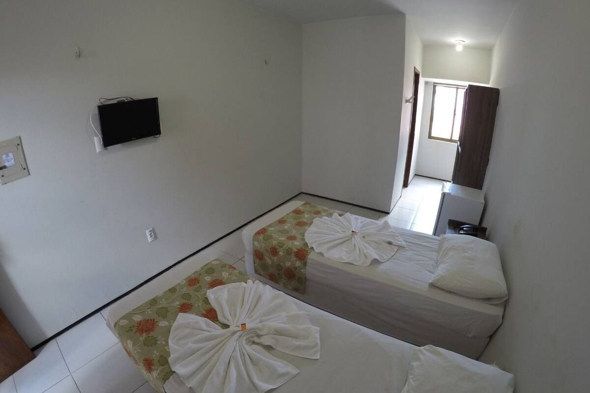 A hotel room with two beds, each with a floral bedspread and white towels. There is a small television on the wall and a window with a curtain.