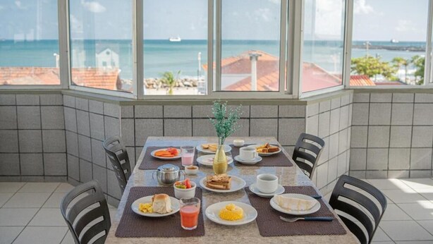 A dining area with a table set for breakfast, large windows with a view of the sea and buildings, and tiled walls. - Iracema Mar Hotel (Fortaleza)