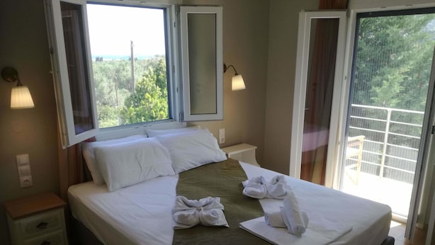 Cama bem arrumada com lençóis brancos e detalhes em marrom, mesa de cabeceira com abajur e vista para o verde através de uma janela.