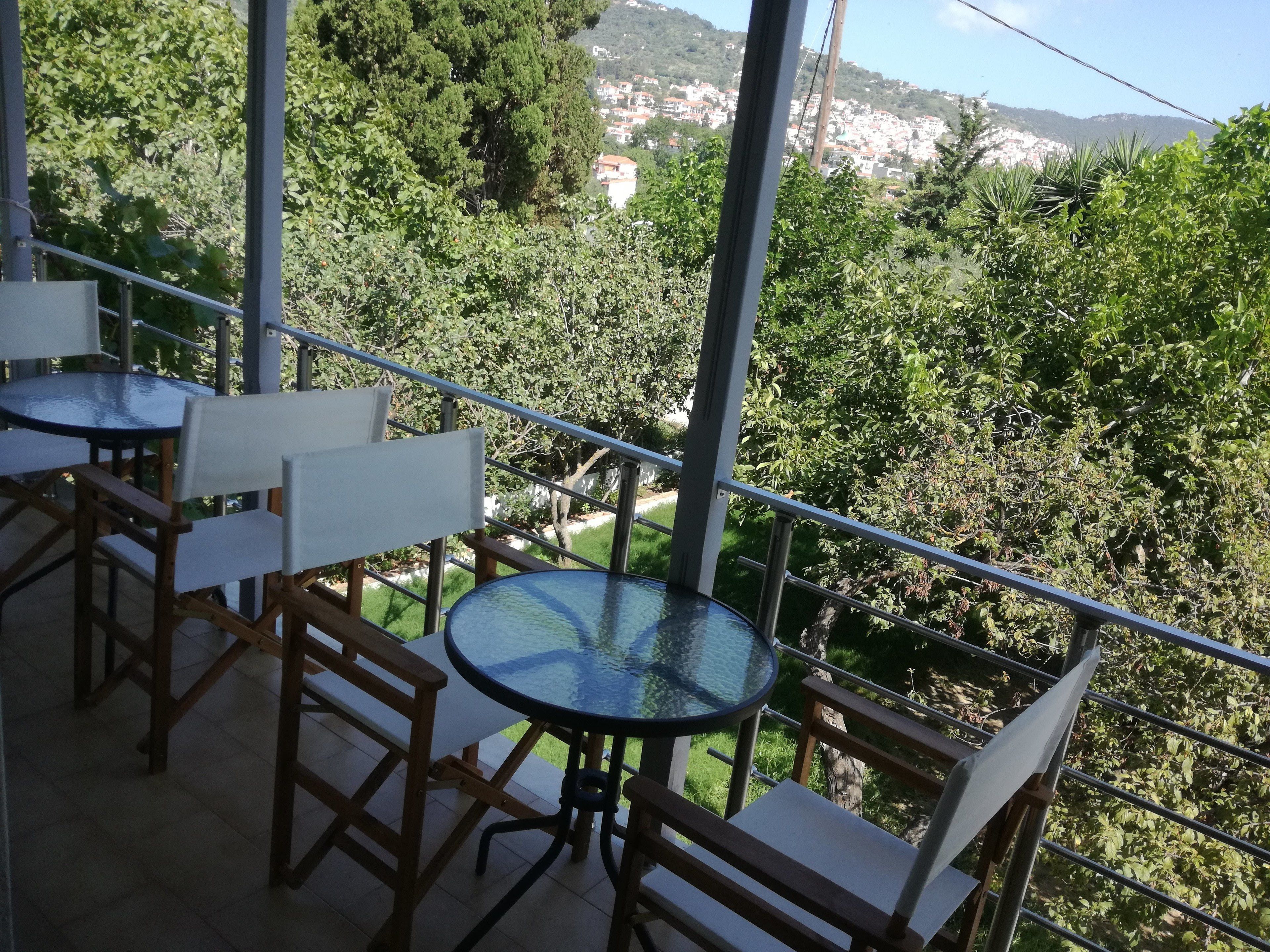 Uma varanda com mesa e cadeiras de vidro, com vista para uma paisagem verdejante e uma cidade distante.