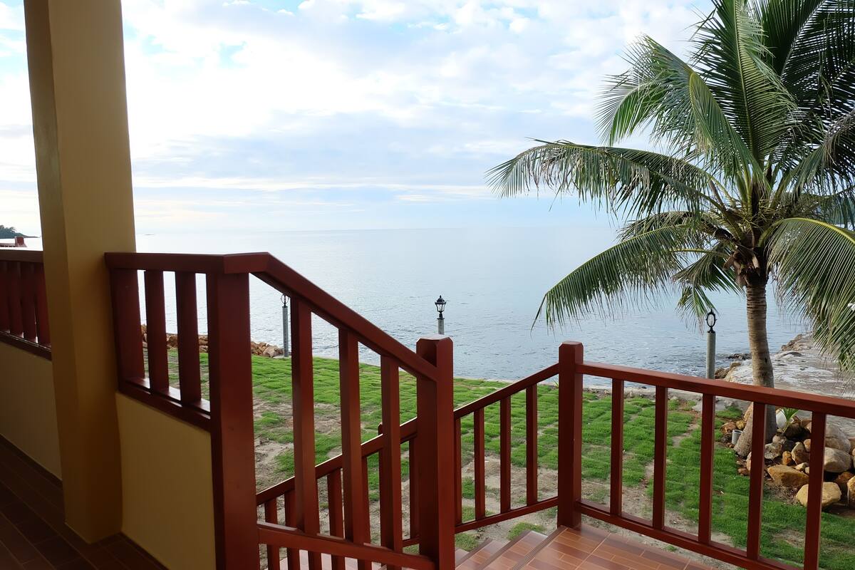 Una escalera de madera que lleva a un balcón con vista a una playa, una palmera y un farol.