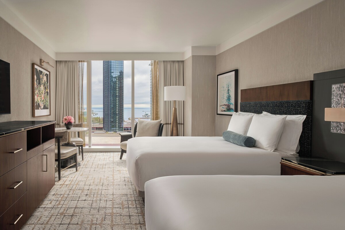 A hotel room with two beds, a desk, a chair, a TV, and a view of a cityscape.