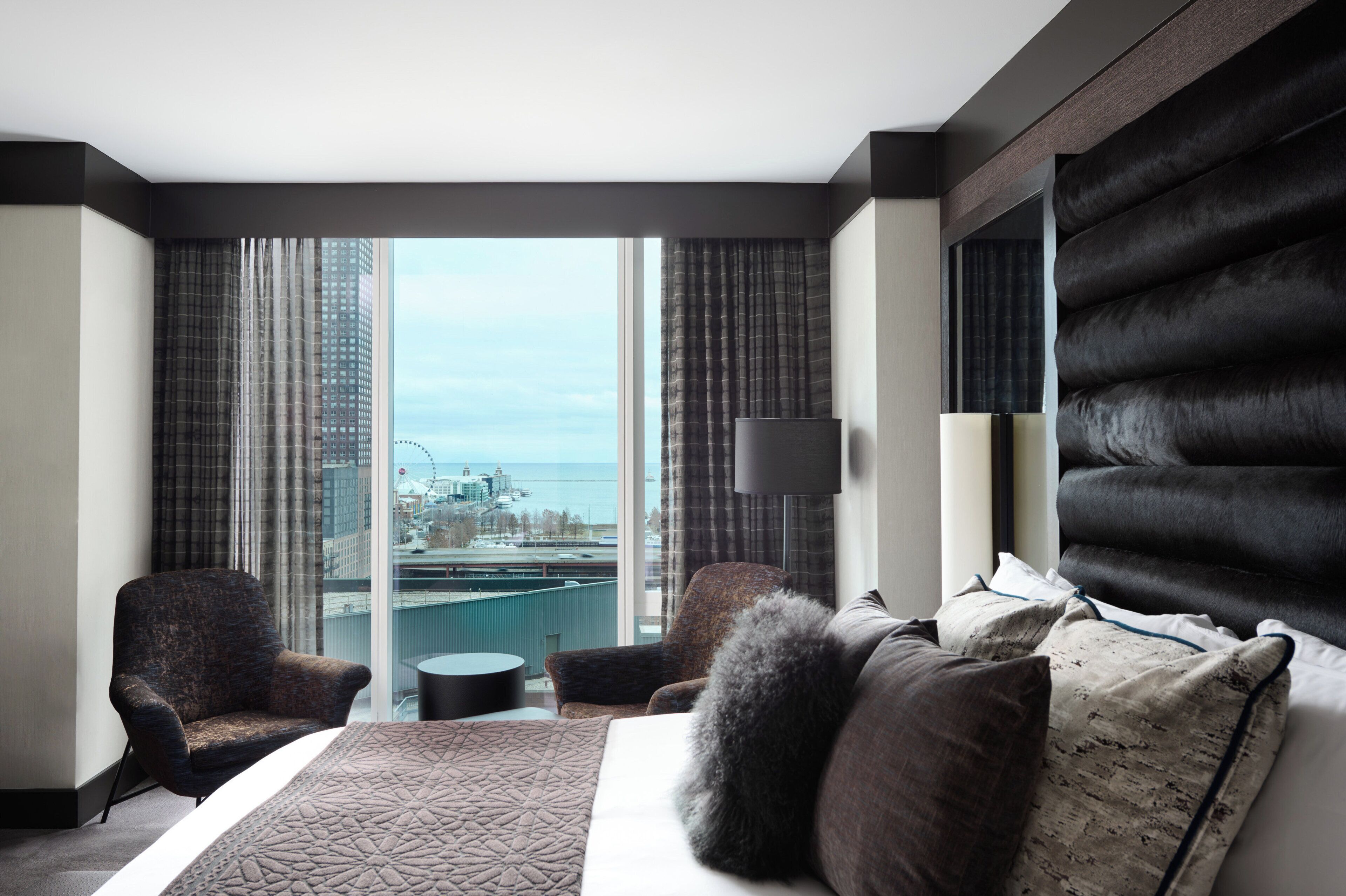 Una habitación de hotel moderna con una cama grande, dos sillones, una mesita redonda y vistas a un paisaje urbano a través de ventanales del suelo al techo.