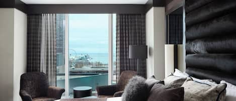 Una habitación de hotel moderna con una cama grande, dos sillones, una mesita redonda y vistas a un paisaje urbano a través de ventanales del suelo al techo.